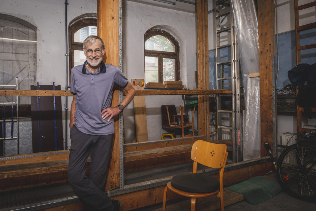 Wolrad Specht trägt ein graues Polohemd und eine dunkelgraue Hose. Er steht in der vorderen linken Hälfte des Bildes. Im Hintergrund ist der Rahmen zu sehen, auf dem er die Teppiche aufspatnnte.