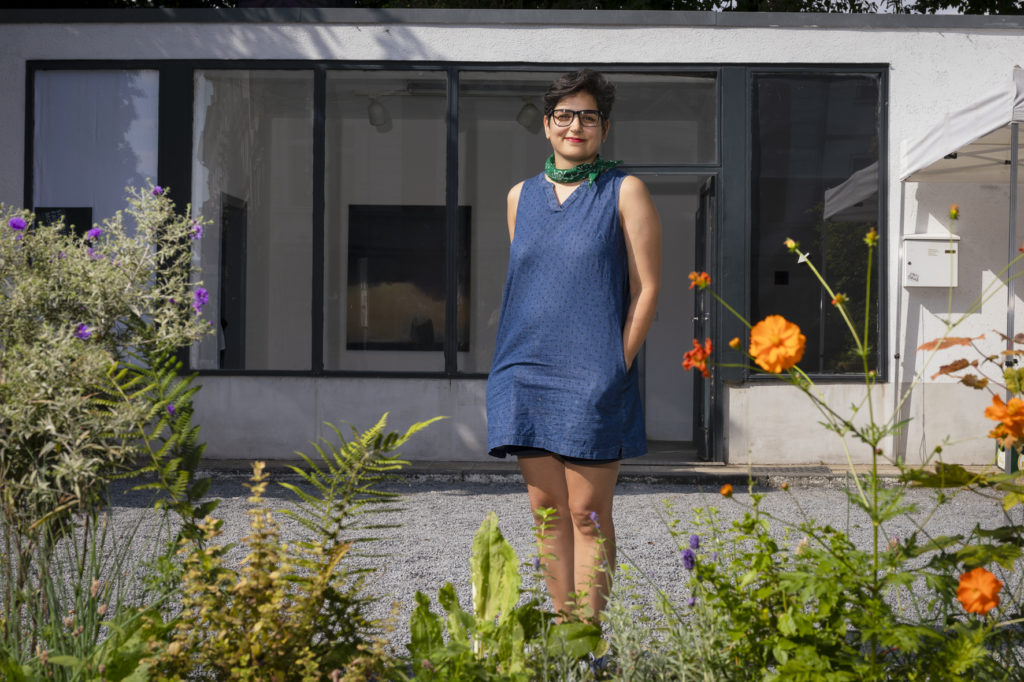 Mona Pourebrahim steht vor der Hebebühne. Im Vordergrund sind Blumen zu sehen. Im Hintergrund ist durch ein Fenster eines von ihren Bildern zu sehen