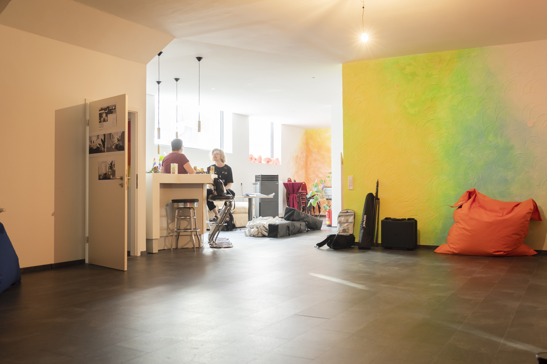 Das Bild zeigt die Räumlichkeiten des Inside:Out. Im Hintergrund sitzen Christiane und Mosche an der Bar und unterhalten sich. Man sieht außerdem eine bunte Wand und eine Fensterfront