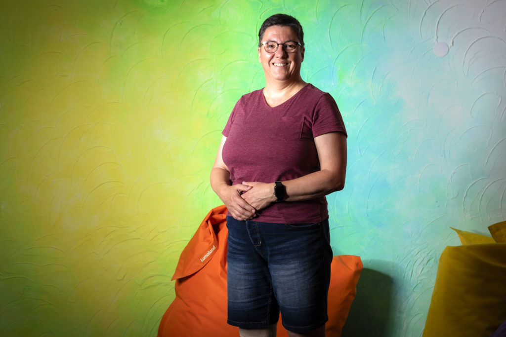 Christiane Freyer hat eine Brille auf, trägt ein weinrotes Tshirt und eine blaue kurze Jeans. Sie steht vor einer bunten Wand und lächelt in die Kamera.