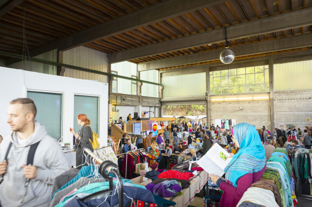 Ein Gewusel von Menschen in einer Industriehalle, die sich durch die engen Gänge des Trödelmarktes drängen. Im Vordergrund ist ein Stand zu sehen der Kleidung verkauft und eine Frau, die in einem Buch liest