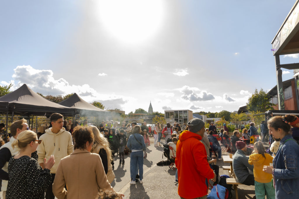Menschen stehen vor der Alten Glaserei und unterhalten sich, während im Hintergrund der blaue Himmel und die Silhouette der Friedhofskirche zu sehen ist