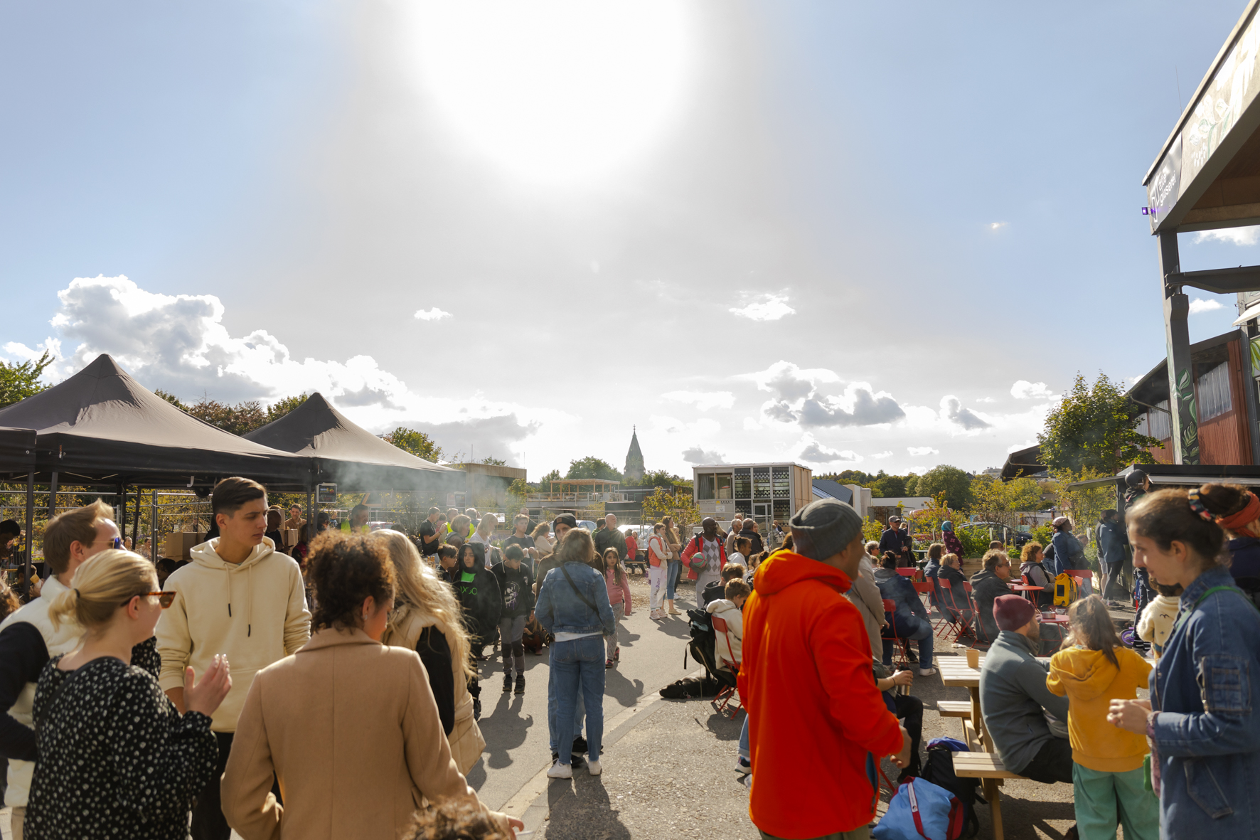 Menschen stehen vor der Alten Glaserei und unterhalten sich, wÃ¤hrend im Hintergrund der blaue Himmel und die Silhouette der Friedhofskirche zu sehen ist